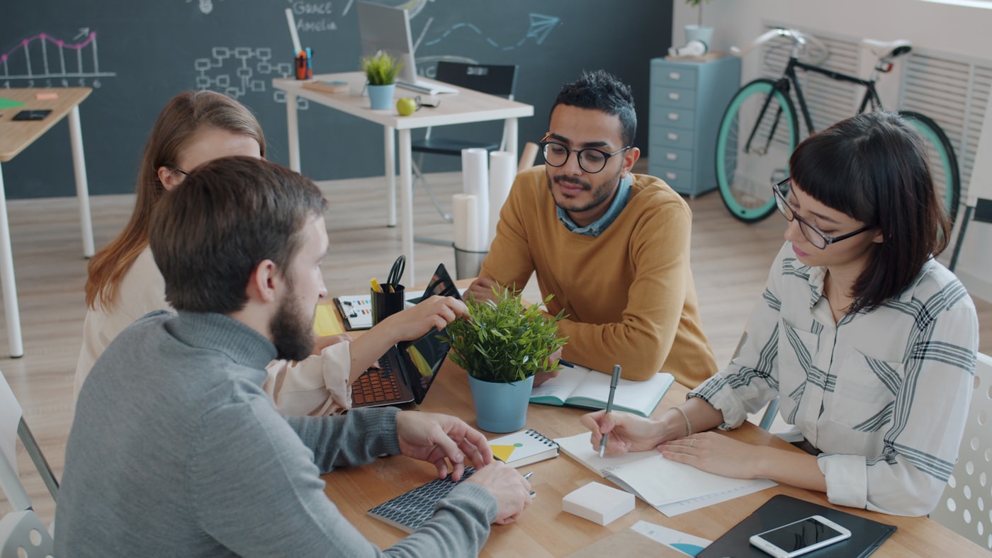 Marketing team collaborating around a desk with a plant, reviewing social and email performance in a bright office